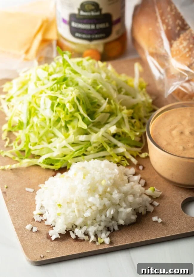 Cutting board displaying neatly diced white onion, shredded iceberg lettuce, and a glass bowl brimming with homemade Big Mac sauce, all prepped for burger assembly.