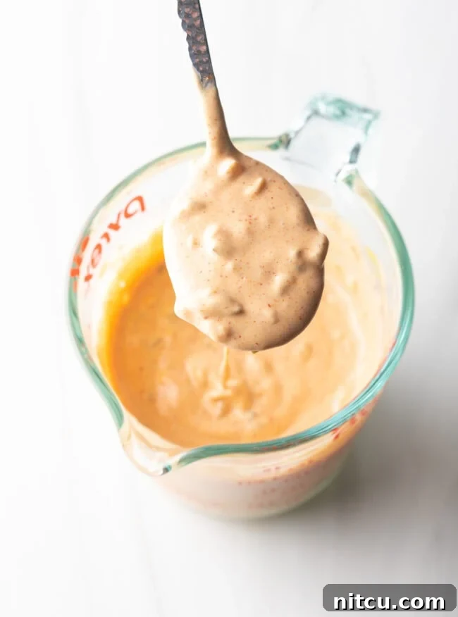 Top-down view of a glass measuring cup filled with freshly made homemade burger sauce, with a spoonful held up to the camera to show its creamy texture.