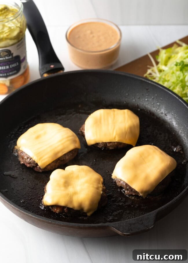 Four sizzling, home-cooked hamburger patties with melted American cheese nestled in a black cast iron skillet, ready for assembly.