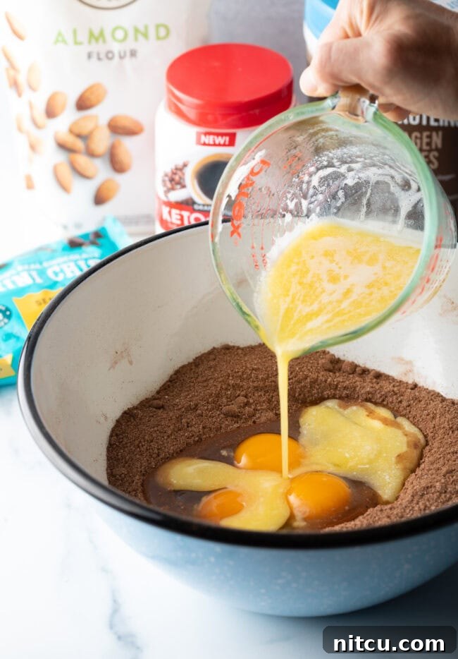 Glow-Up Protein Brownies 7 A hand pouring melted butter from a glass measuring cup into a bowl containing the dry ingredients and eggs, beginning the mixing process.
