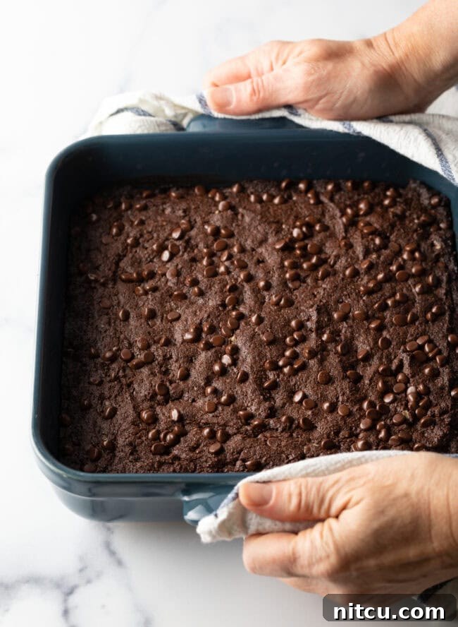 Glow-Up Protein Brownies 9 A hand holding a square pan of freshly baked brownies, dotted with melted chocolate chips, just out of the oven.