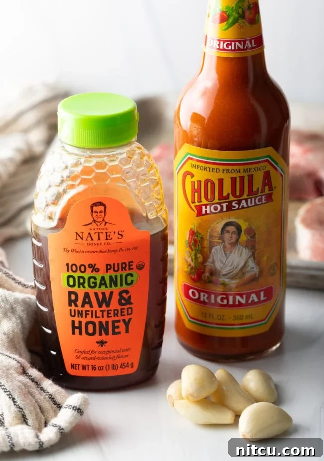 A simple arrangement of Cholula hot sauce, a jar of raw honey, and fresh garlic cloves – the core ingredients for the spicy honey garlic glaze.