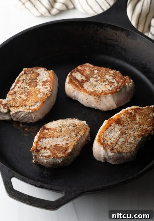 Four seared pork chops, beautifully browned, resting in a cast iron skillet before the glaze is added.