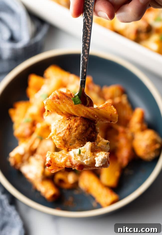 One-Pan Meatball Casserole 12 Dark gray plate with a generous portion of baked meatball casserole with pasta and cheese. Fork is holding a bite to camera.