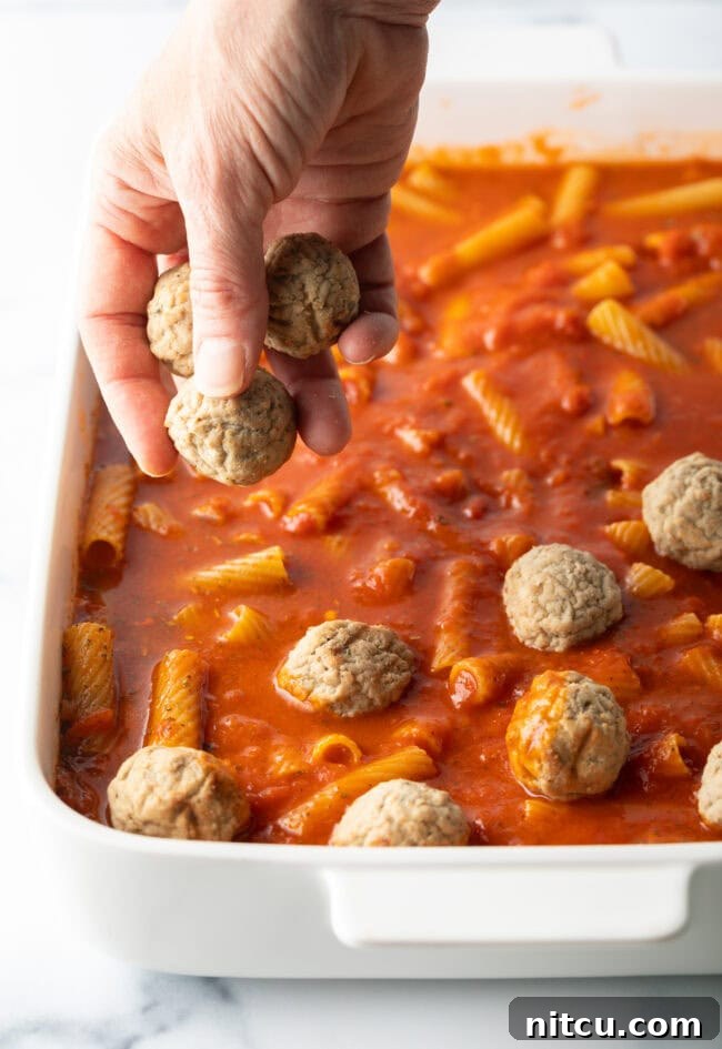 One-Pan Meatball Casserole 6 Hand adding frozen meatballs to baking dish with marinara and pasta.