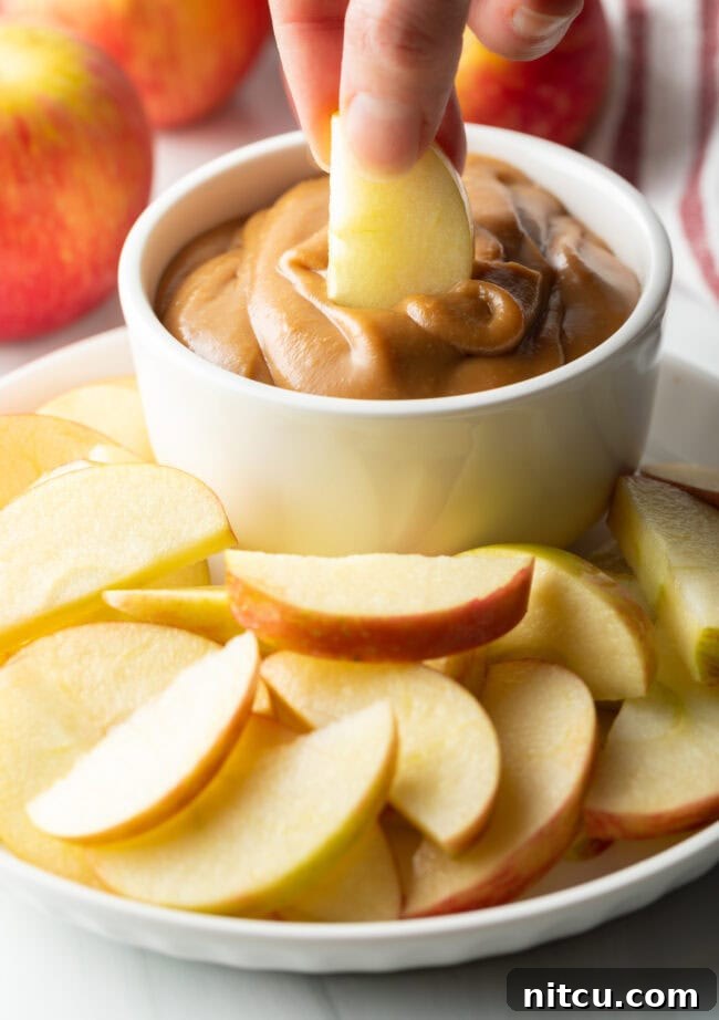 White bowl of cream cheese caramel apple dip, with a piece of apple slice beautifully emerging from the dip, all presented on a white plate amidst more apple slices.