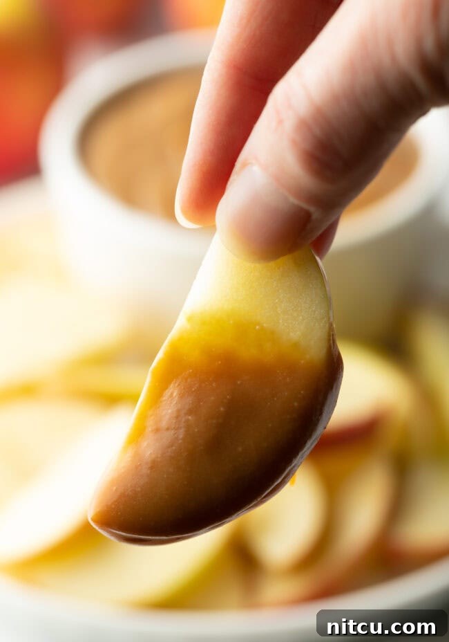 A hand holds an apple slice generously coated with rich caramel dip, presented enticingly towards the camera.