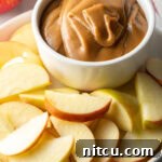 White bowl of cream cheese caramel apple dip, on a white plate surrounded by apple slices.