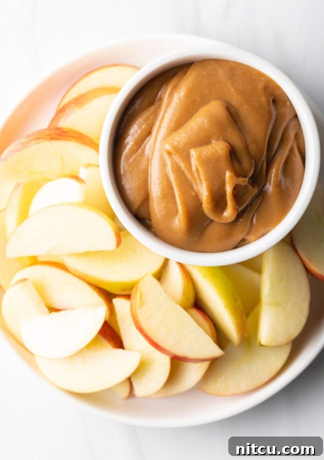 Top-down view of a pristine white bowl filled with creamy caramel apple dip, artfully arranged on a white plate and surrounded by freshly sliced apples.