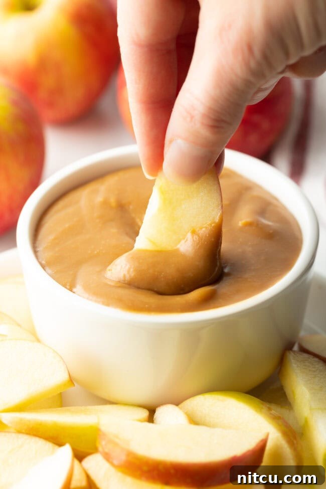 A hand expertly uses a fresh apple slice to scoop a generous portion of the creamy caramel dip from a pristine white bowl, showcasing its perfect dipping consistency.