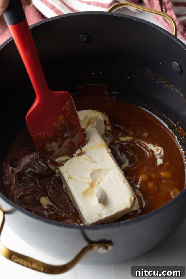 Softened cream cheese being gently added to the warm, cooled caramel mixture in a pot, preparing for whisking.