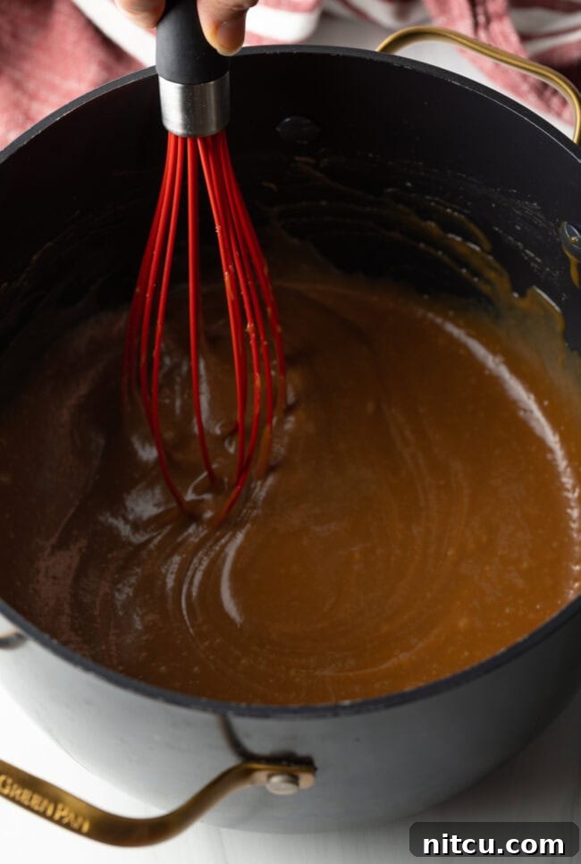 A whisk vigorously blending the caramel cream cheese mixture in a pot until it achieves a perfectly thick, smooth, and lump-free consistency.