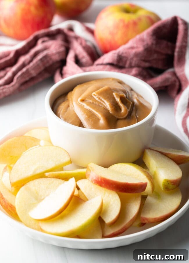 White bowl of creamy caramel apple dip, artfully arranged on a white plate and surrounded by freshly sliced apples.