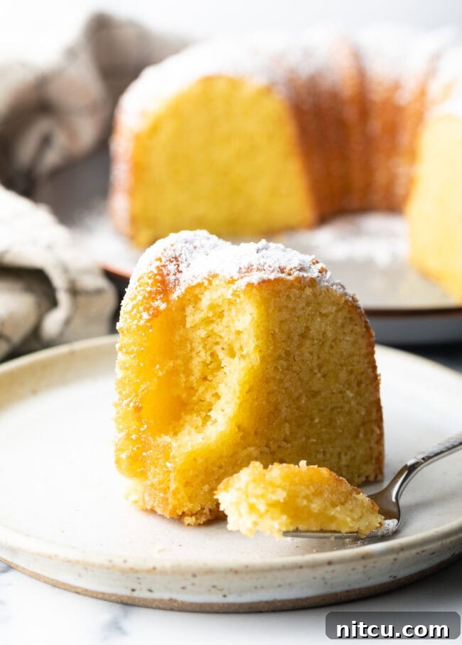Sun-Kissed Lemon Olive Oil Cake 11 A close-up shot of a single slice of moist yellow cake on a white plate, with a fork gently cutting into a bite, highlighting its soft texture and inviting crumb.