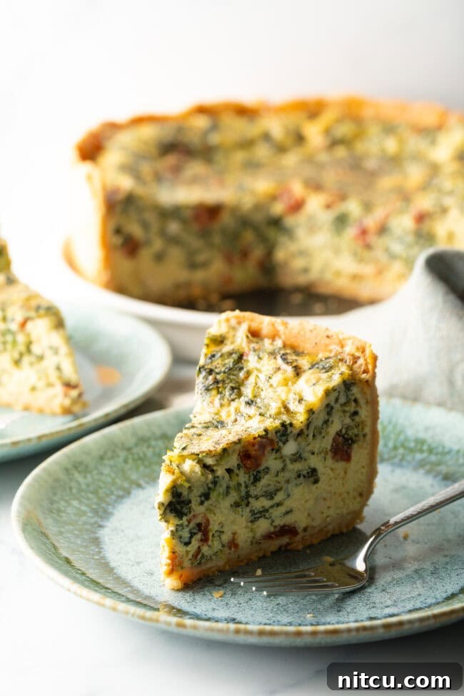 Garden Medley Deep Dish Quiche 12 A close-up of a tall, perfect slice of quiche on a light blue plate, with a fork beside it, highlighting its flaky crust and rich, varied filling.