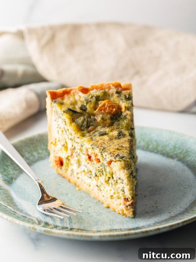 Garden Medley Deep Dish Quiche 13 A delectable slice of quiche, tall and inviting, rests on a light blue plate, accompanied by a fork, ready for enjoyment.