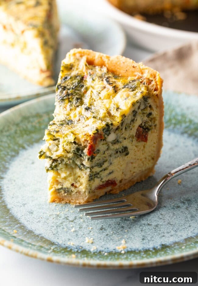 Garden Medley Deep Dish Quiche 14 A tall, perfectly baked slice of quiche on a light blue plate, with a small bite taken from its tip and a fork resting nearby.