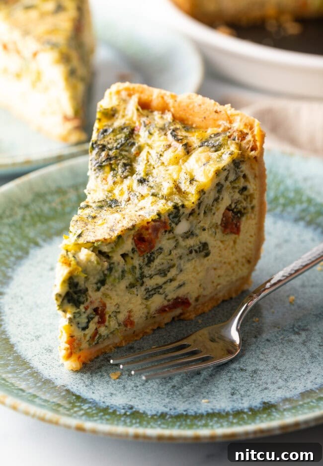 Garden Medley Deep Dish Quiche 4 A tall, perfectly baked slice of vegetarian quiche rests on a light blue plate, with a fork positioned invitingly beside it. The visible layers showcase the flaky crust and the abundant vegetable filling.