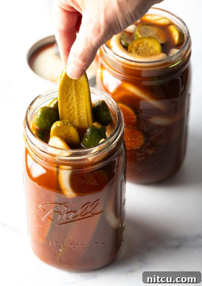 Chamoy Drenched Pickles 12 Two large mason jars of chamoy pickles. The tops are off of the jars, and a long pickle is being pulled out of one jar.