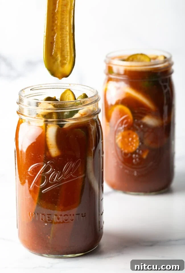 Chamoy Drenched Pickles 3 Two large mason jars of chamoy pickles. The tops are off of the jars, and a long pickle is being pulled out of one jar.