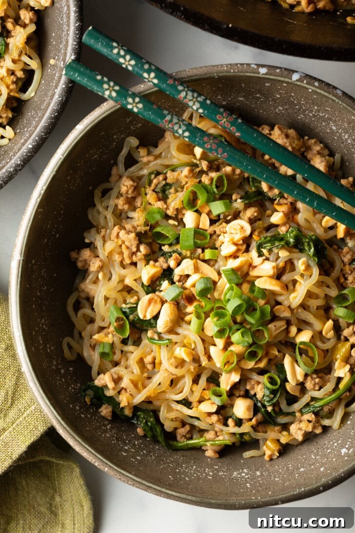 Spicy Szechuan noodles in a black bowl topped with green onion slices. Set of chopsticks are resting on top of the bowl.