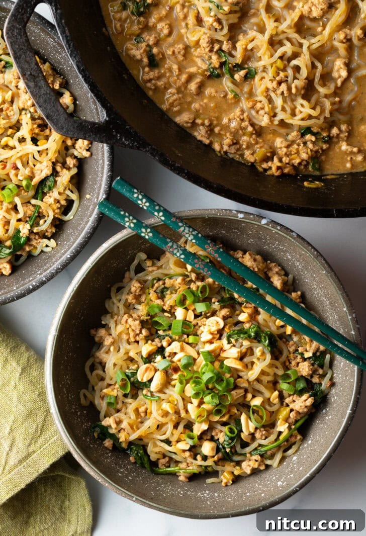 Spicy Szechuan noodles in a black bowl topped with green onion slices. Set of chopsticks are resting on top of the bowl.