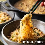 Spicy Szechuan noodles in a black bowl topped with green onion slices. Hand with set of chopsticks are grabbing a bite of noodle.