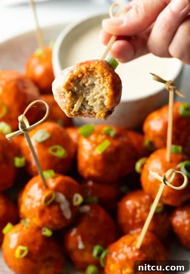 Plate of buffalo chicken meatballs skewered with serving toothpicks, nestled next to a white dish of ranch dressing dipping sauce. A hand is holding a meatball by the toothpick, showing a bitten inside to camera.