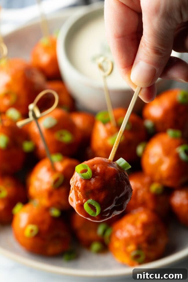 Plate of buffalo chicken meatballs skewered with serving toothpicks, nestled next to a white dish of ranch dressing dipping sauce. A hand is holding a meatball by the toothpick, showing to camera.