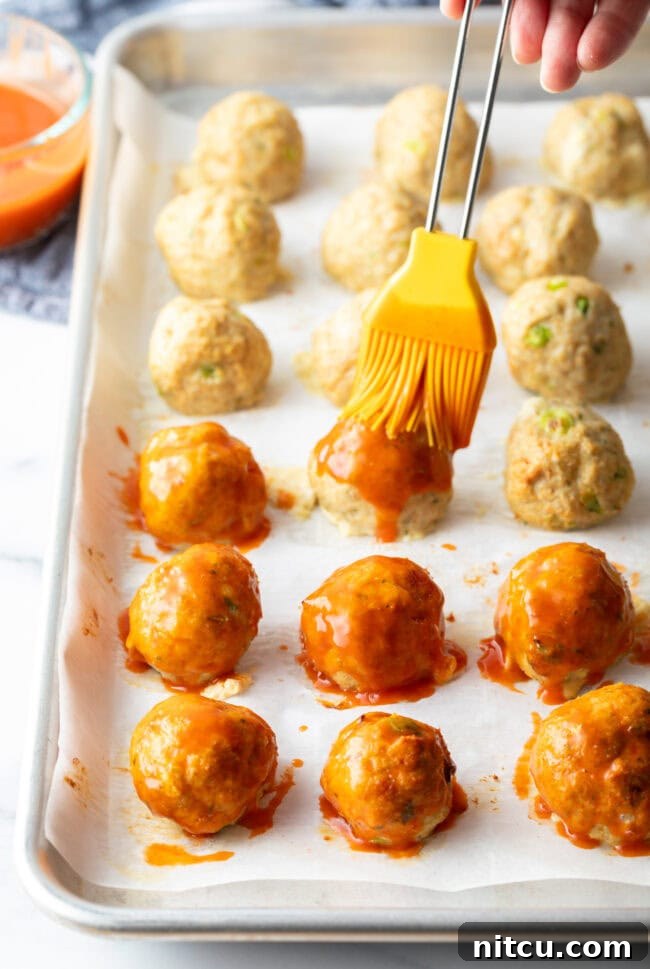 A dozen meatballs on a baking sheet, yellow basting brush adding buffalo sauce to the meatballs.