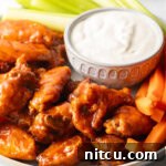 Plate of saucy buffalo chicken wings. On the plate is also a ramekin of ranch dressing, a pile of carrot sticks, and a pile of celery sticks.