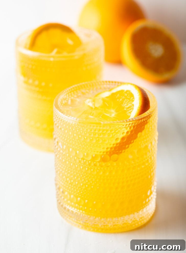 Two orange crush cocktails in glasses, each garnished with a slice of orange. There are fresh oranges and lemons around the glasses.