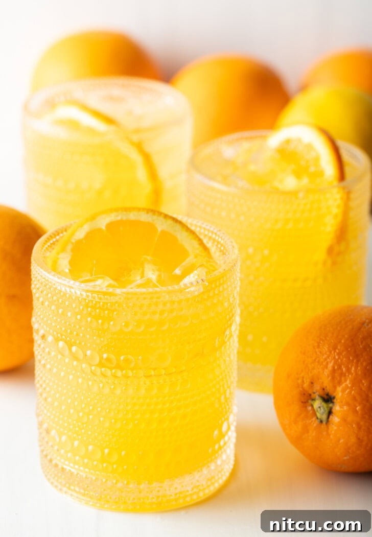 Two orange crush drinks in glasses, each garnished with a slice of orange. There are fresh oranges and lemons around the glasses.