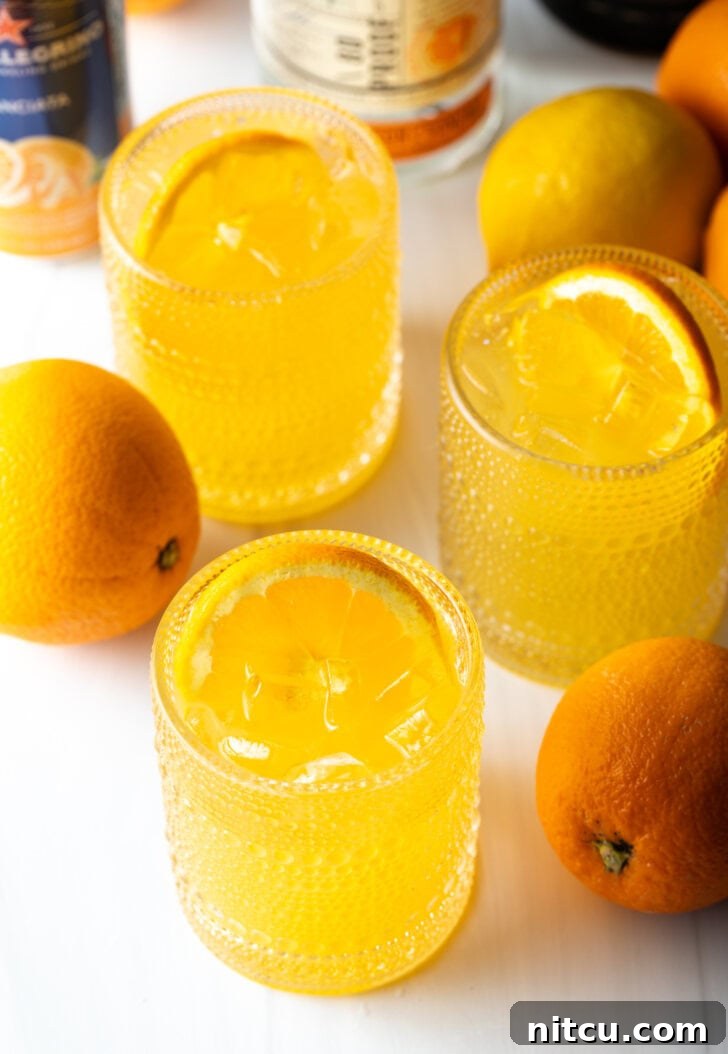 Three orange crush cocktails in glasses, each garnished with a slice of orange. There are fresh oranges and lemons around the glasses.