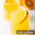 Two orange crush cocktails in glasses, each garnished with a slice of orange. There are fresh oranges and lemons around the glasses.