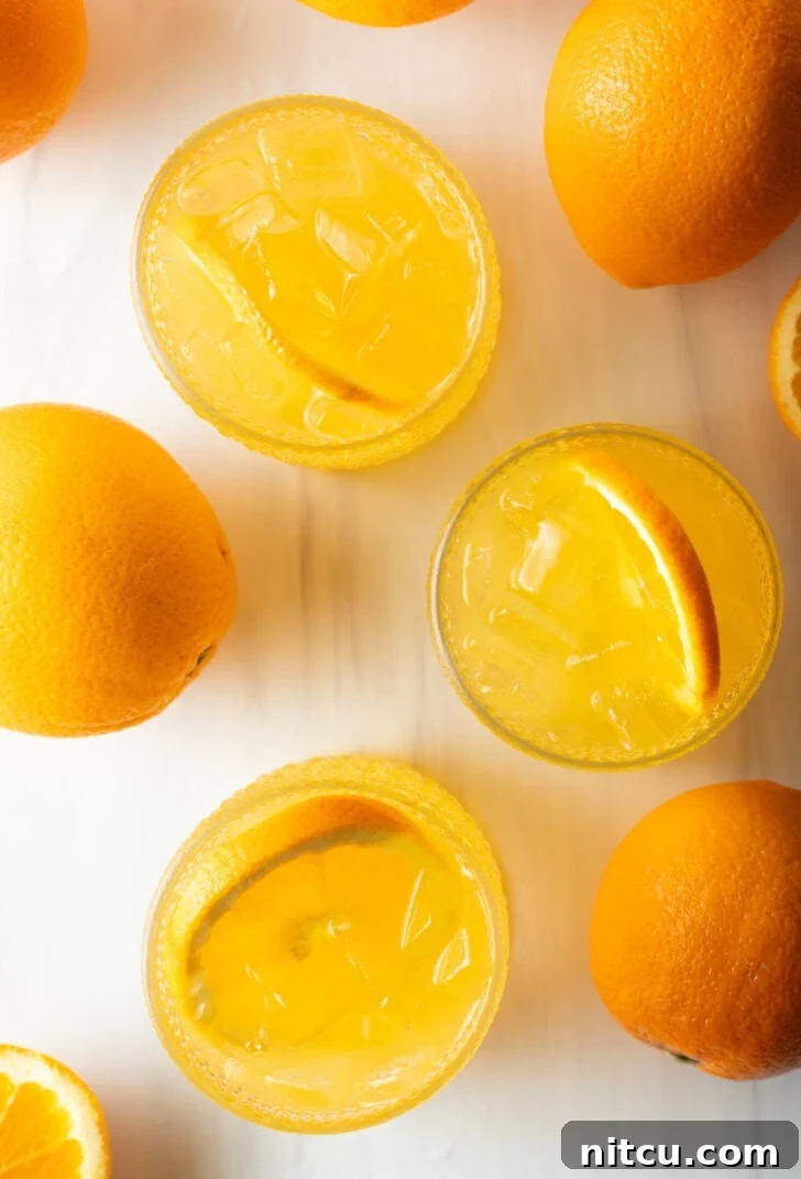 Top down view 4 orange crush drinks in glasses, each garnished with a slice of orange. There are fresh oranges and lemons around the glasses.
