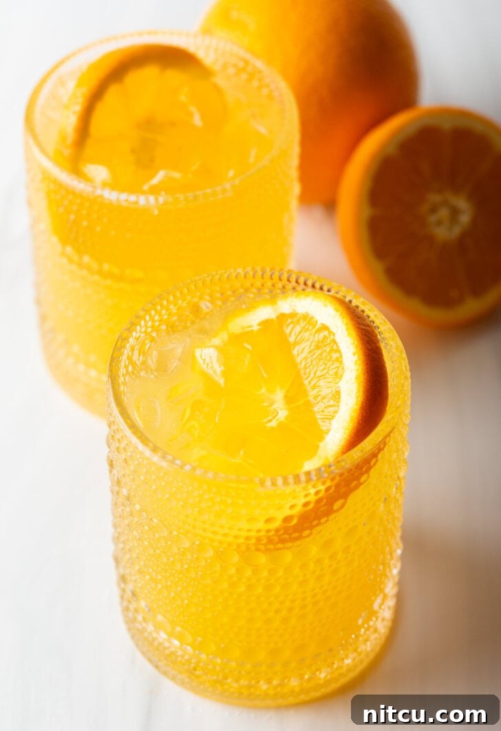 Two orange crush cocktails in glasses, each garnished with a slice of orange. There are fresh oranges and lemons around the glasses.