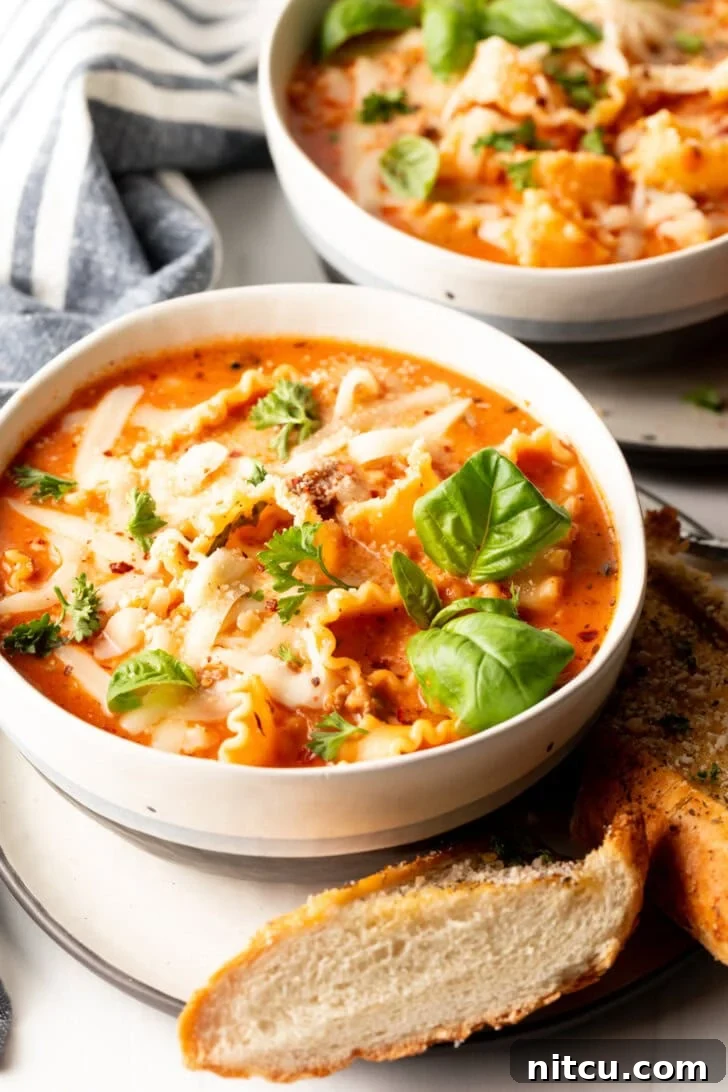 One-Pot Lasagna Delight 2 A vibrant white bowl overflowing with homemade lasagna soup, beautifully garnished with two fresh basil leaves. A rustic slice of baguette rests on a white plate beneath the bowl, ready for dipping.