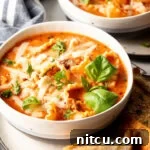 A white bowl loaded with lasagna soup, garnished with two fresh basil leaves. A slice of baguette is on a white plate under the bowl.