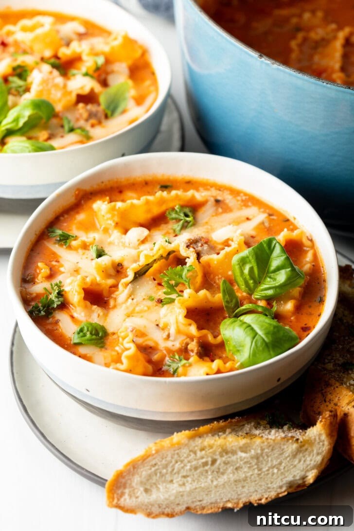 One-Pot Lasagna Delight 10 A inviting white bowl of lasagna soup, garnished with vibrant fresh basil leaves, with a side of toasted baguette on a white plate, ready to be enjoyed.