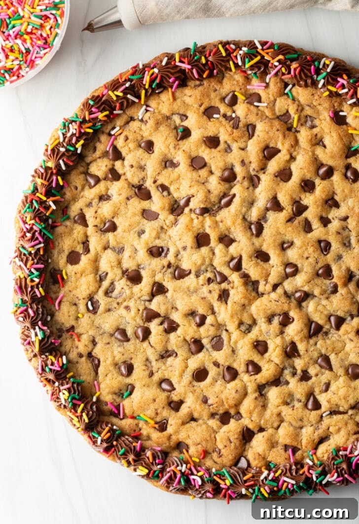 Ultimate Chocolate Chip Cookie Cake 2 Top down view large chocolate chip cookie cake. The outside edge is piped with chocolate frosting and covered with rainbow sprinkles.