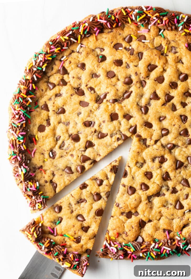 Ultimate Chocolate Chip Cookie Cake 12 Large chocolate chip cookie cake. The outside edge is piped with chocolate frosting and covered with rainbow sprinkles. A cake cutter is taking a wedge slice from the cake.