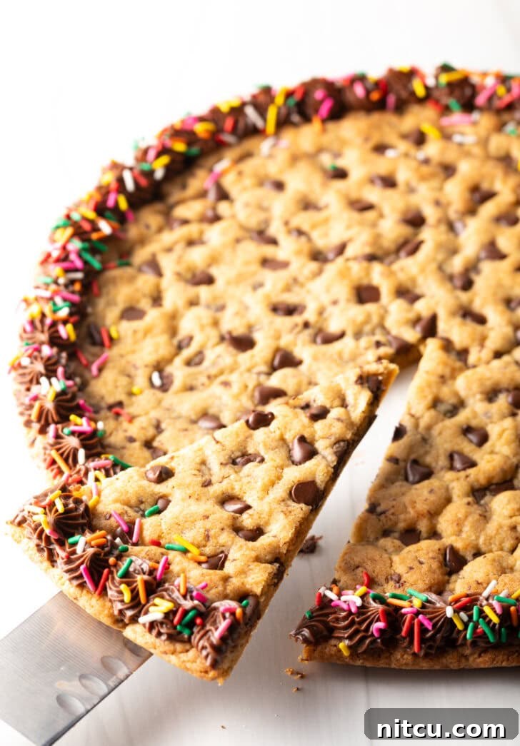 Ultimate Chocolate Chip Cookie Cake 3 Large chocolate chip cookie cake. The outside edge is piped with chocolate frosting and covered with rainbow sprinkles. A cake cutter is taking a wedge slice from the cake.