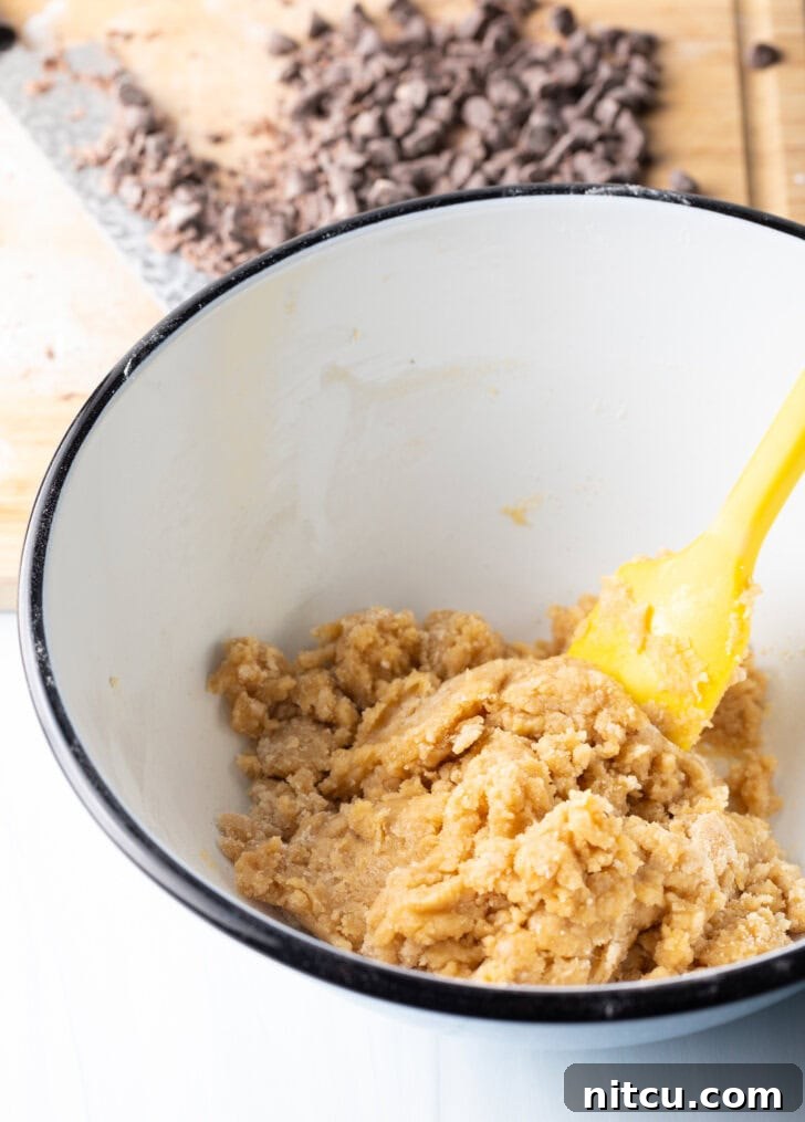 Ultimate Chocolate Chip Cookie Cake 6 White mixing bowl with cookie dough ingredients, stirred by a yellow spatula.