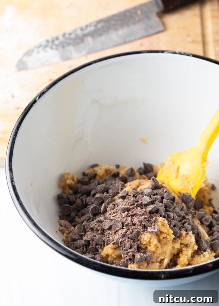 Ultimate Chocolate Chip Cookie Cake 7 White mixing bowl with cookie dough ingredients, stirred by a yellow spatula.