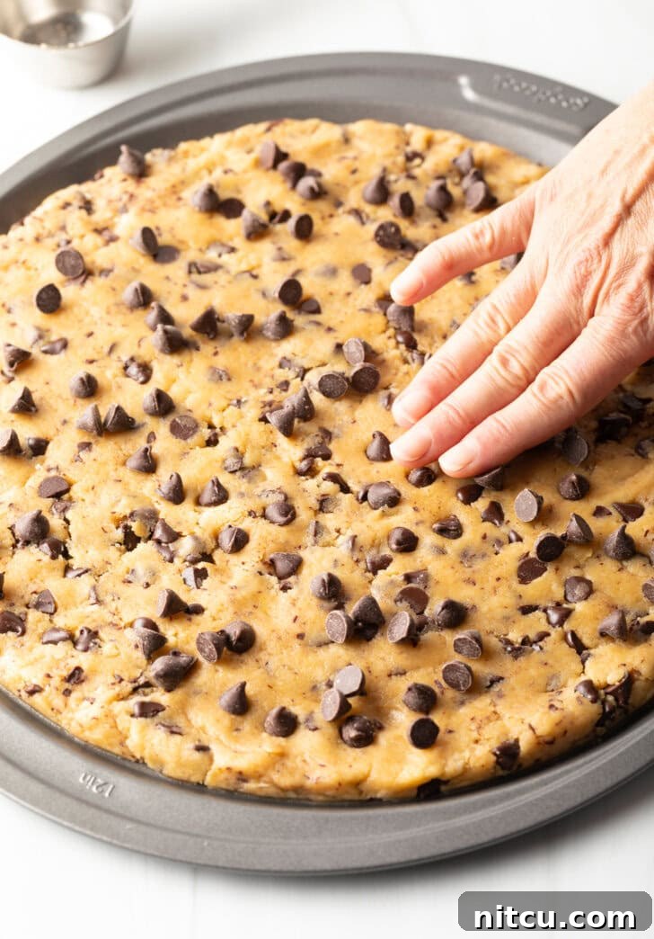 Ultimate Chocolate Chip Cookie Cake 8 Large chocolate chip cookie cake, hand spreading dough out in a pizza pan.