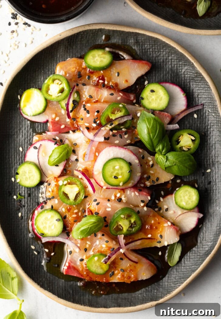 Top down view of black oval plate loaded with thin slices of yellowtail crudo topped with seasonings, thin slides of radishes, jalapenos, red onion, and a sprinkle of sesame seeds.
