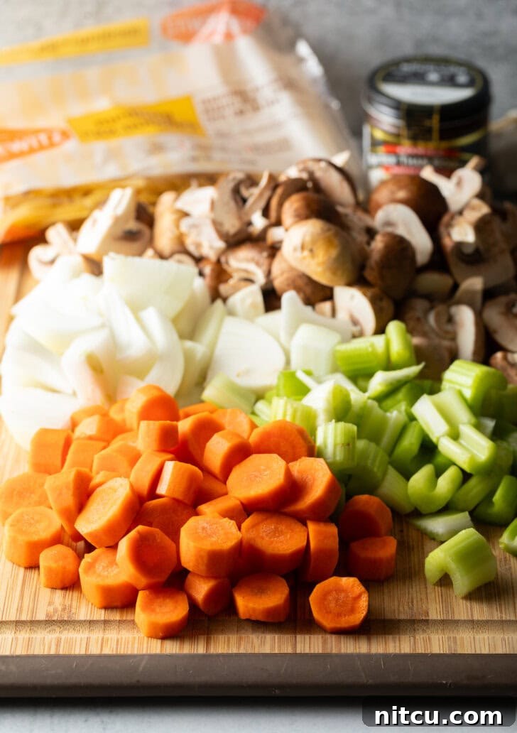 Ultimate Beef Noodle Soup 4 Soup ingredients, chopped in individual piles on a wood cutting board are a pile of carrots, onions, celery, and mushrooms.