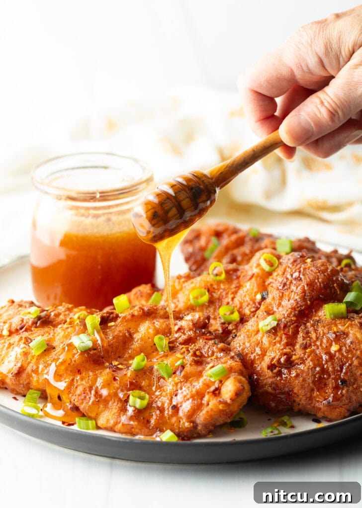 Plate of fried chicken tenders with small glass jar of hot honey to the side, A hand with a wooden honey spoon is drizzling honey over the breaded fried tenders.