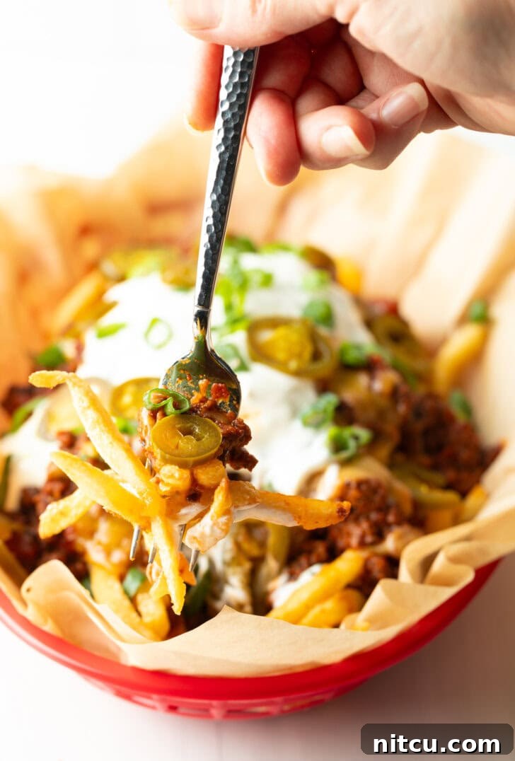 Smothered Chili Cheese Fries 2 A rustic basket lined with parchment paper, overflowing with golden, crispy french fries. They are generously smothered with a rich, hearty chili, and topped with a glistening layer of melted cheddar cheese. A dollop of cool sour cream, a scattering of fresh chopped green onions, and vibrant sliced jalapeños complete the presentation. A hand holding a forkful of this delicious creation is visible, ready to enjoy.